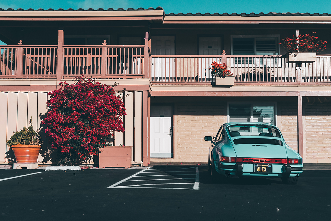 Teal vintage Porsche 911 parked in a motel-style courtyard with a pinkish building, wooden balcony, potted plants, and a large red flowering bush.