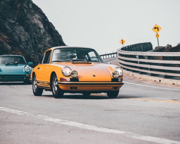 Wall decor photo of classic orange Porsche 911 on winding coastal road followed by a turquoise Porsche, perfect for vintage automotive interior decoration