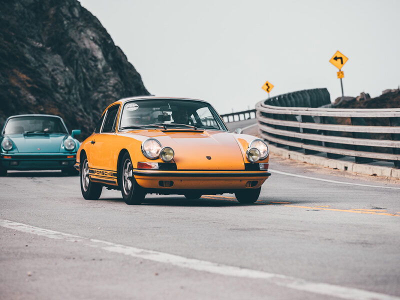 Wall decor photo of classic orange Porsche 911 on winding coastal road followed by a turquoise Porsche, perfect for vintage automotive interior decoration