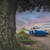 Photo Porsche GT3 RS et Sunset Premium automotive photograph of a blue Porsche GT3 RS parked on a peaceful country road at dusk, framed by a majestic tree with rolling hills in the background
