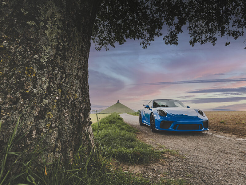 Premium automotive photograph of a blue Porsche GT3 RS parked on a peaceful country road at dusk, framed by a majestic tree with rolling hills in the background