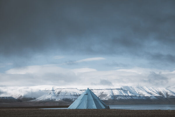 Photo Svalbard tente Fjord Svalbard photography showing a solitary blue tipi tent facing snow-capped mountains of the arctic fjords under a dramatic sky