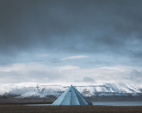 Svalbard photography showing a solitary blue tipi tent facing snow-capped mountains of the arctic fjords under a dramatic sky