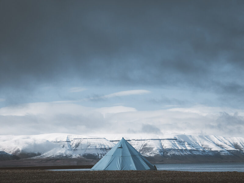 Svalbard photography showing a solitary blue tipi tent facing snow-capped mountains of the arctic fjords under a dramatic sky