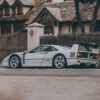 Photographie Ferrari F40 Grey Ferrari F40 supercar in profile view with iconic rear wing and side air intakes, photographed against rustic European architecture with wooden fence and traditional buildings