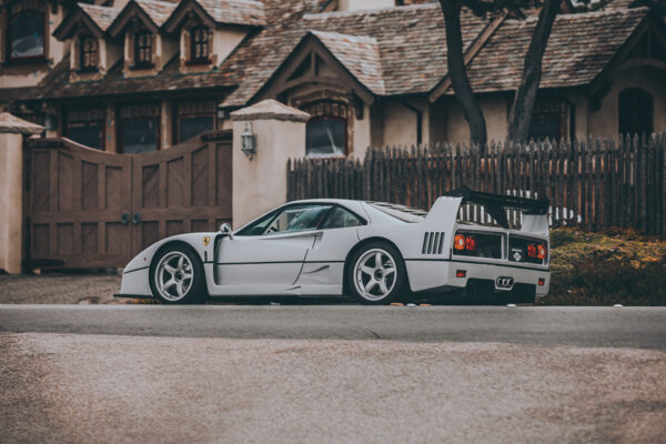 Photographie Ferrari F40 Grey Ferrari F40 supercar in profile view with iconic rear wing and side air intakes, photographed against rustic European architecture with wooden fence and traditional buildings