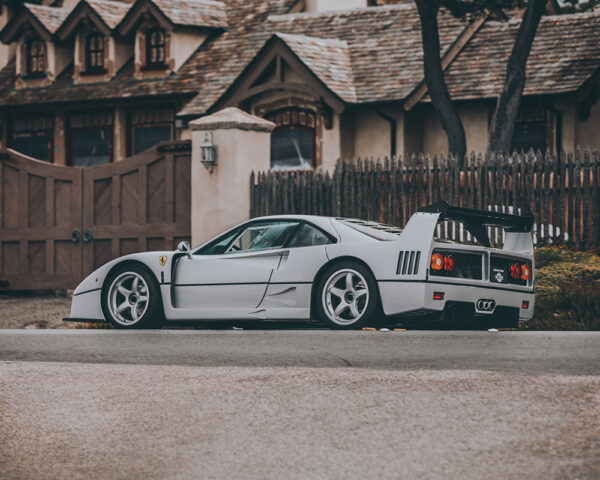 Grey Ferrari F40 supercar in profile view with iconic rear wing and side air intakes, photographed against rustic European architecture with wooden fence and traditional buildings