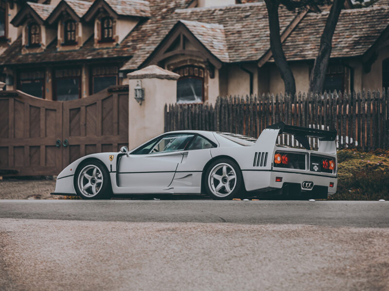 Grey Ferrari F40 supercar in profile view with iconic rear wing and side air intakes, photographed against rustic European architecture with wooden fence and traditional buildings