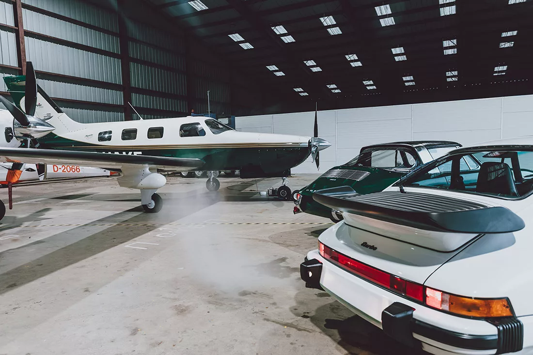 White Porsche 911 Turbo and green 911 in front of a Piper Malibu in a hangar lit by skylights - fine art photography print