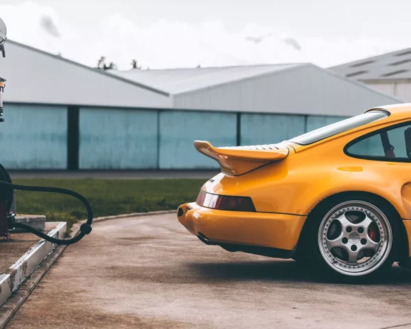 Yellow Porsche 930 Turbo S fine art photography print, rear three-quarter view beside a vintage red fuel pump and corrugated hangar, Cars and Roses