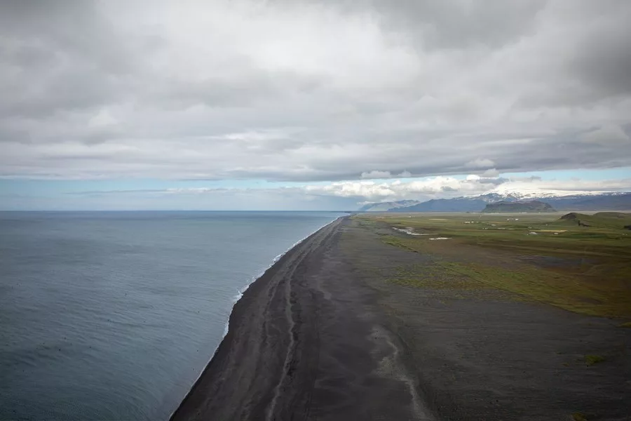 Black sand beach on the coast of Iceland seen from above, with the grey Atlantic, green plains and snow-capped mountains