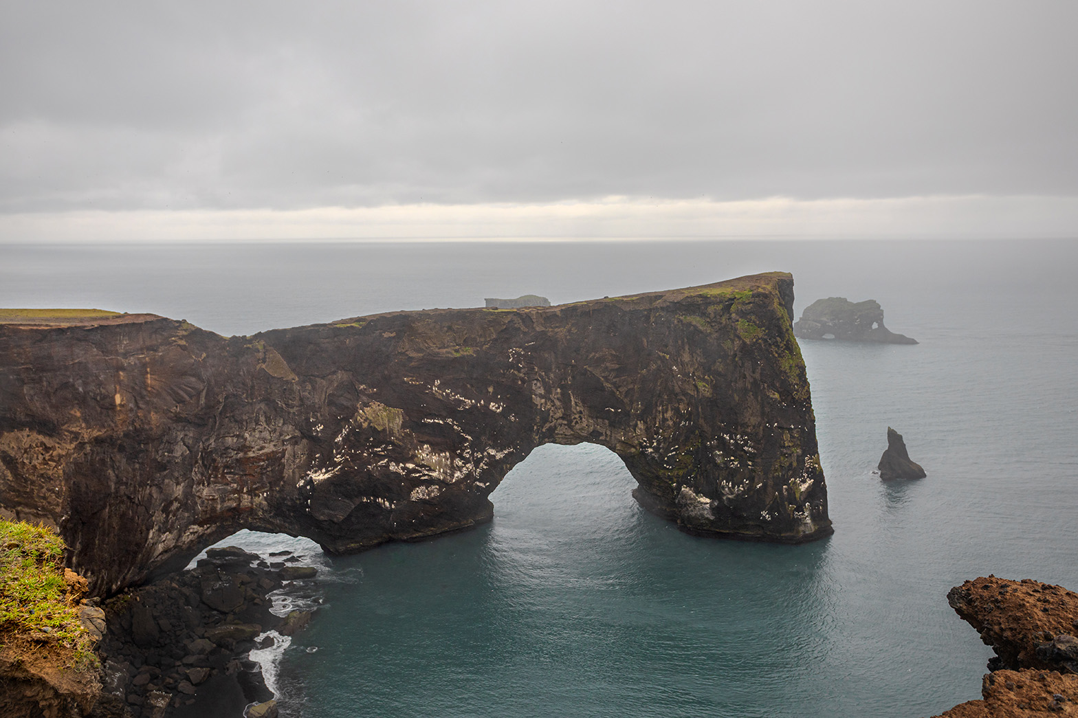 Majestic coastal arch over calm blue-green waters, rugged cliffs, and distant rock formations under cloudy skies for landscape photography wall art.