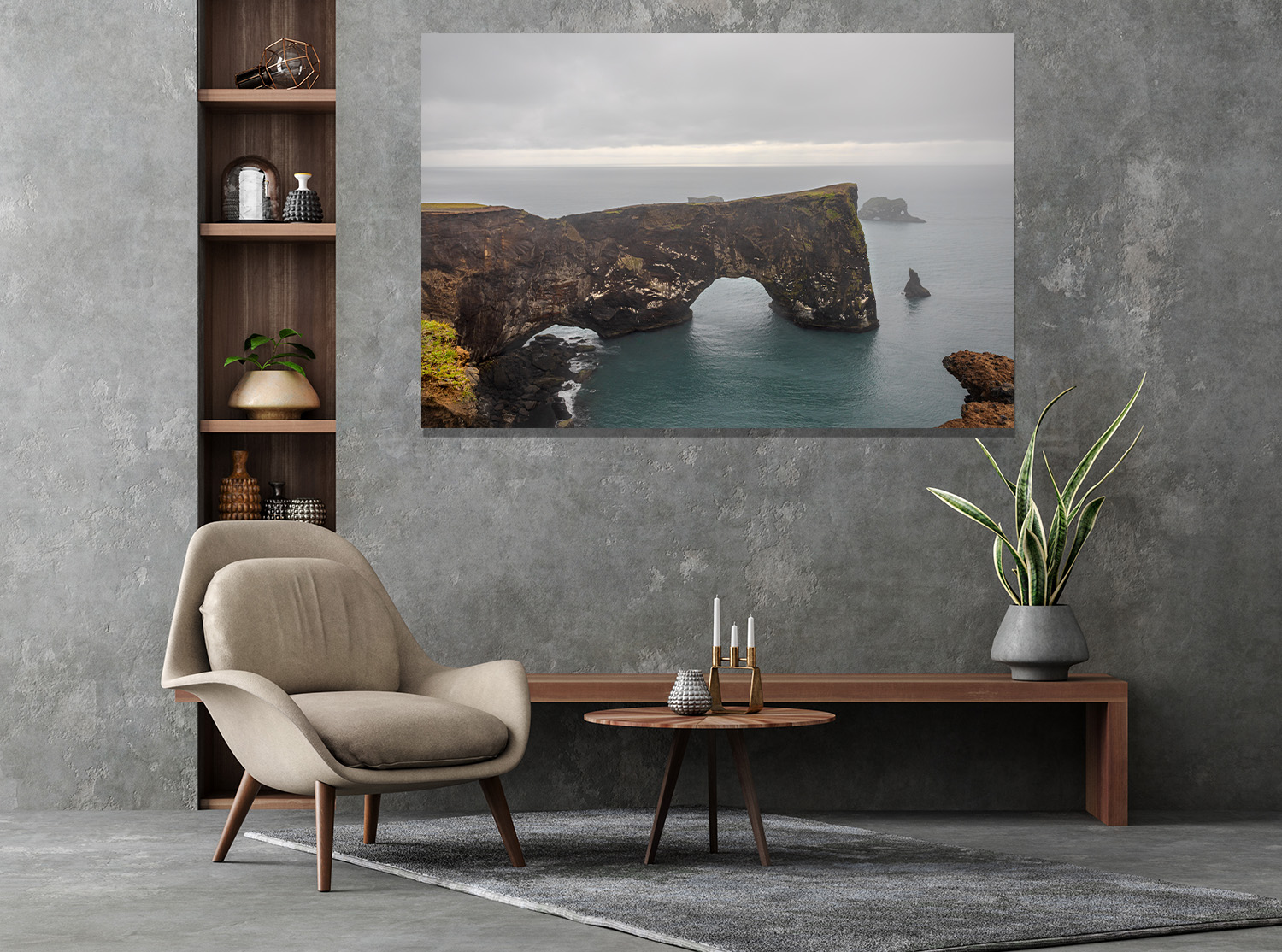 A modern living room with a large framed landscape photo of a coastal rock arch on the gray textured wall, flanked by a wooden shelving unit, a beige armchair, a round wooden coffee table, a long wooden console, a potted snake plant, and a gray rug.